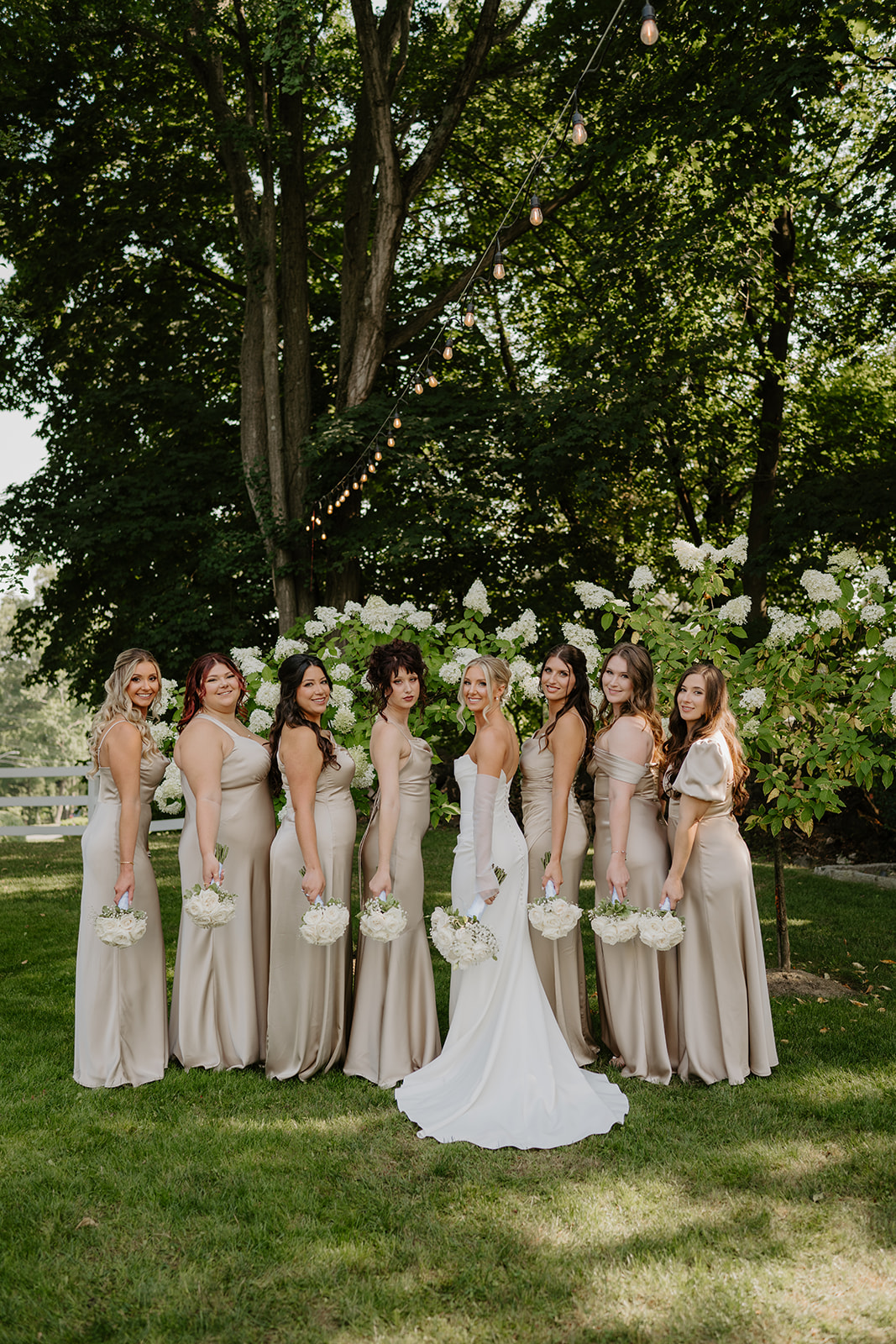 Bride and bridesmaids in champagne satin dresses stand in a line on the lawn, holding white bouquets.